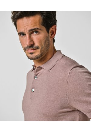 Homme aux cheveux courts et foncés et à la barbe, portant un polo mauve, regardant confiamment vers la caméra sur un fond uni.