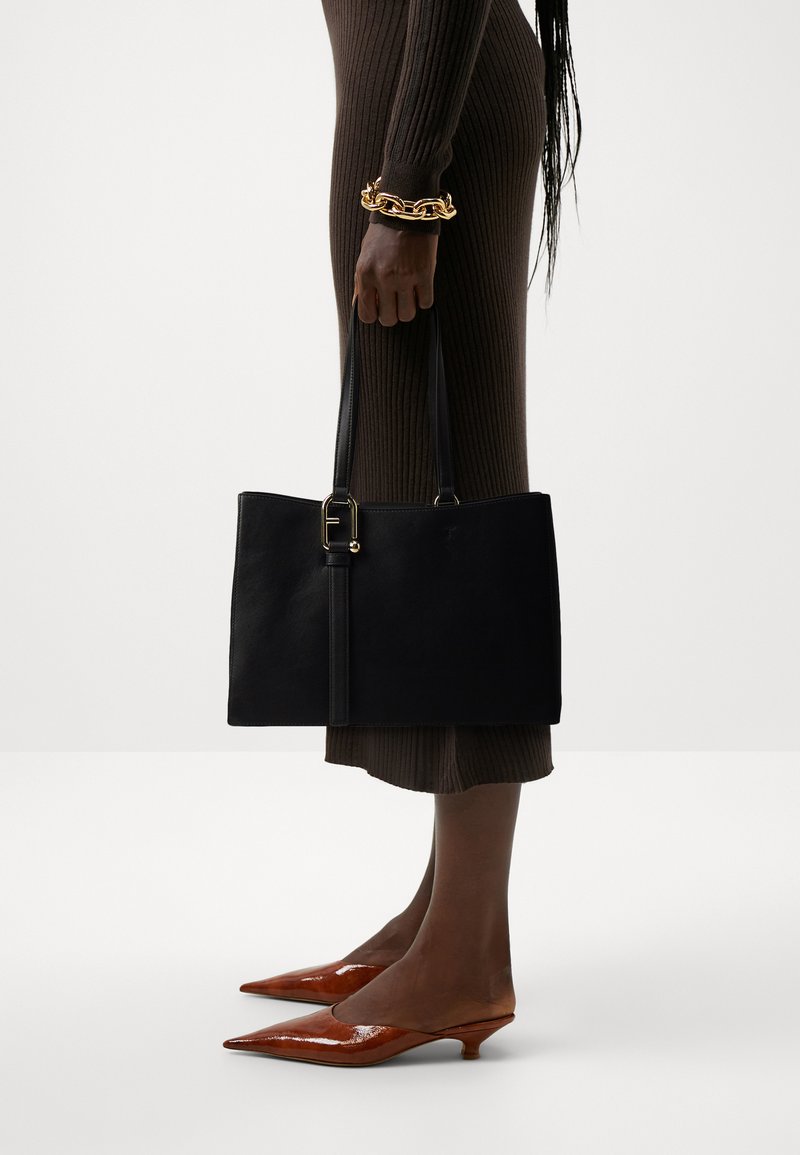 Sac fourre-tout en cuir noir avec double anses, ornée d'un accent doré et d'une forme structurée. Porté avec une robe marron à côtes et des talons bruns pointus.
