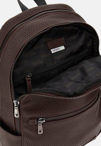 Zaino in pelle sintetica marrone con texture, dotato di due tasche con zip, interno foderato nero e logo 'GUESS' su un'etichetta. Accenti in metallo argentato visibili.