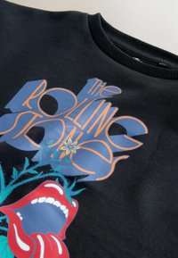 Sort sweatshirt med dristigt grafisk design, der indeholder røde læber og blå blomsterelementer, som viser teksten "The Rolling Stones" i orange.
