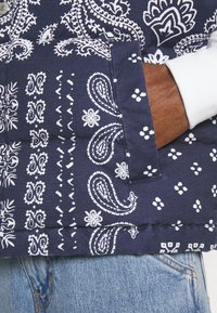 Marinblå puffväst med vita paisley- och blommönster, med en sidoficka och en vit innerfoder. Denimjeans syns.