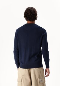Tmavě modrý pletený svetr s kulatým výstřihem, žebrovanými manžetami a lemem. Nesený přes béžové cargo kalhoty, které ukazují hladkou texturu.
