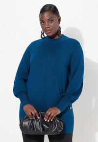 Maglione azzurro lavorato a maglia con maniche lunghe a palloncino, polsini a coste e orlo a onda. Il modello tiene una borsa a tracolla nera in pelle. Materiali lisci e testurizzati.
