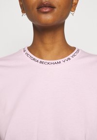 Světle růžové tričko s kulatým výstřihem, které má černý logový pruh s nápisem "VVB VICTORIA BECKHAM" kolem límce. Měkká bavlněná struktura.