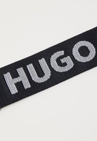 Sangle en tissu noir avec le mot "HUGO" brodé en lettres majuscules blanches.