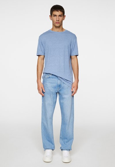 Camiseta de algodón azul claro con cuello redondo, combinada con jeans rectos de lavado claro y zapatillas blancas, de pie sobre un fondo liso.