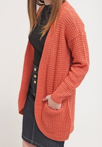 Cardigan corallo a maglia aperta con maniche lunghe, vestibilità comoda e tasche anteriori. Realizzato con filato testurizzato e orli a coste.