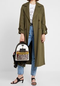 Persona con abrigo trench color verde oliva, camiseta gráfica blanca, jeans azul claro, tacones negros, sosteniendo una mochila blanca con estampado de leopardo.