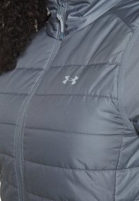 Gray padded vest with a zippered front, featuring light gray logo. Smooth texture with horizontal quilted pattern and hood.