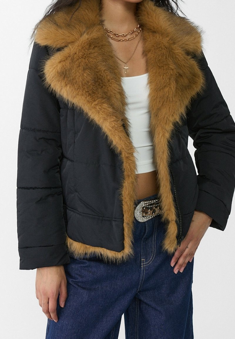 Femme portant une doudoune noire avec une bordure en fausse fourrure brune, un crop top blanc, un jean bleu, des colliers en couches en or et une boucle de ceinture argentée décorative.