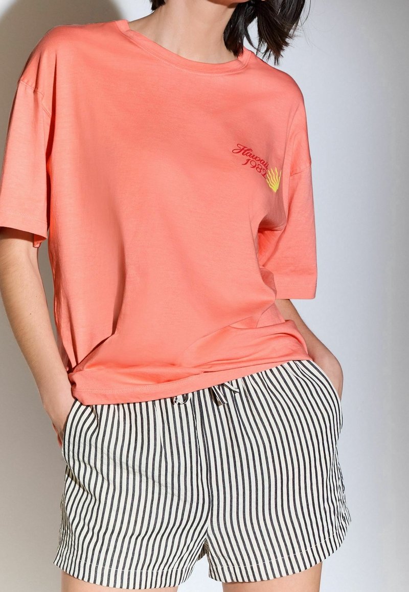 Camiseta de gran tamaño de color coral con mangas cortas, con texto y diseño en color amarillo. Combinada con pantalones cortos a rayas en blanco y negro, con cintura elástica.