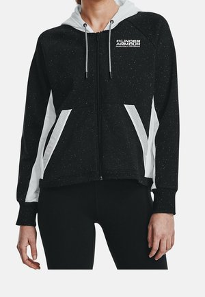 Frau trägt einen schwarzen gesprenkelten Kapuzenpullover mit Reißverschluss, weißen Mesh-Einsätzen, einer weißen Kapuze und dem Under Armour-Logo auf der linken Brust.