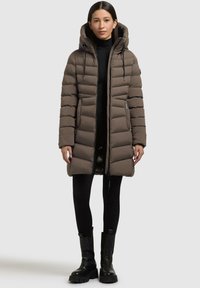 Giubbotto imbottito marrone con cappuccio, dotato di chiusura con zip, texture trapuntata, maniche lunghe e tasche laterali. Indossato sopra un outfit nero.
