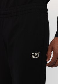 Sorte joggebukser lavet af blødt materiale, med elastisk talje, sidelommer og et lille EA7-logo i lyse farver på venstre lår.