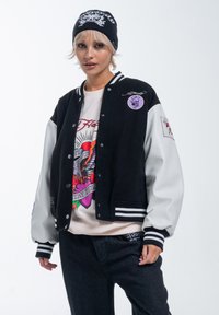 Black and white varsity jacket featuring leather sleeves, side stripes, and embroidered patches. Worn over a graphic sweater and dark jeans.