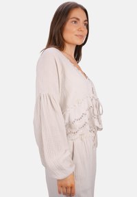 Beige verkürztes Oberteil aus strukturiertem Stoff, mit Spitze akzentuiert, V-Ausschnitt und langen puffartigen Ärmeln, die an der Taille gebunden sind. Die dazu passenden Hosen sind nicht sichtbar.