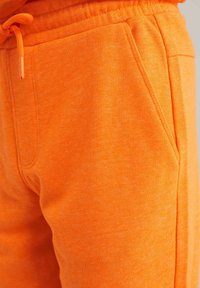 Oranžové tepláky s jemnou textúrou, elastický pás s šnúrkou a bočnými vreckami. Materiál sa zdá byť hladký a neformálny.