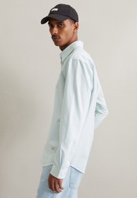 Lichtblauwe en witte gestreepte overhemd met lange mouwen, met knoopmanchetten en een zijlabel, in combinatie met lichtdenim jeans en een zwarte pet.