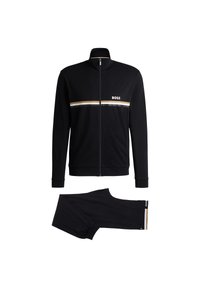 Schwarzer Trainingsanzug bestehend aus einer Jacke mit Reißverschluss, die einen weißen und goldenen horizontalen Streifen sowie ein Logo aufweist, kombiniert mit passenden schmal zulaufenden Hosen.
