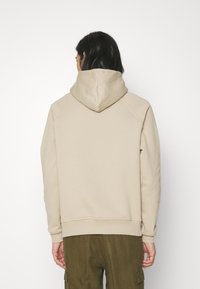 Béžová mikina s velkou kapucí, žebrovanými manžetami a lemem. Má hladkou texturu a raglánové rukávy. Nošena s olivově zelenými kalhotami.