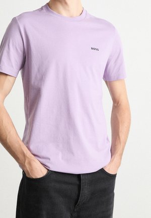 Mand iført en lys lilla BOSS T-shirt med korte ærmer og sorte bukser med hænderne i lommerne mod en ensfarvet baggrund.