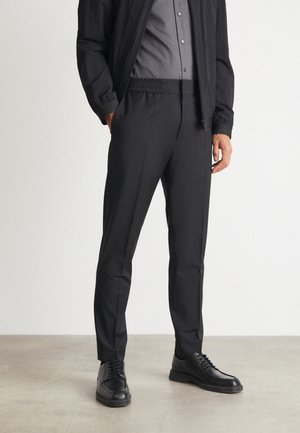Mand iført sorte slim fit bukser, sort casual jakke, grå skjorte med knapper og sorte snørebåndssko, stående på en grå gulv.