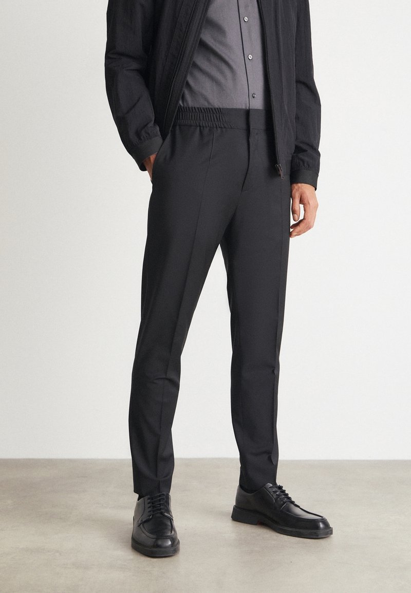 Homme portant un pantalon slim noir, une veste décontractée noire, une chemise grise boutonnée et des chaussures noires à lacets, debout sur un sol gris.