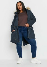 Marinblå parka-jacka med fuskpäls på huvan, dragkedja och knäppningar, framfickor och dragsko i midjan. Matchad med blå jeans och vita sneakers.