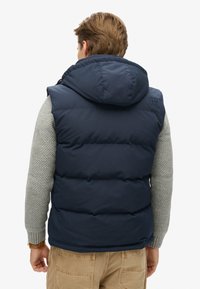 Navy blue puffer vest with a hood, featuring a quilted texture. Worn over a light gray knitted sweater, with a zipper and adjustable hood.