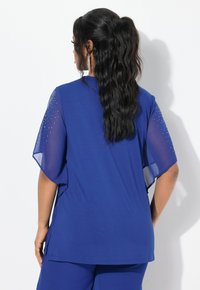 Top blu con maniche corte e trasparenti decorate con strass. Il tessuto è liscio e leggero, con una silhouette rilassata.