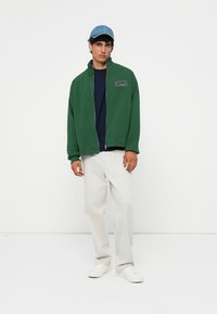 Groene zip-up jack met hoge kraag, donkerblauw t-shirt, beige broek en witte sneakers, aangevuld met een lichtblauwe pet.