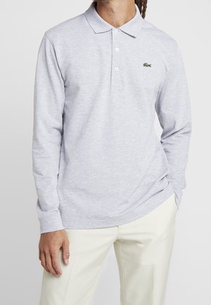 Mand iført lysegrå langærmet polo skjorte med tre knapper og lille grønt krokodillelogo på brystet, kombineret med off-white bukser.