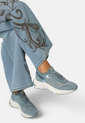 Blauwe sneakers met mesh- en suèdetail, gedragen met lichtblauwe jeans met bruine borduursels van bloemenpatronen op brede pijpen.