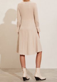 Robe en tricot côtelé beige avec des manches longues et une jupe évasée. Présente un corsage ajusté et une texture lisse, associée à des bottines blanches.
