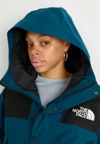 Tirkizna vodootporna jakna s crnim akcentima, veliki kapuljača i zip džepovi. Ima bijeli logo The North Face na prsima.