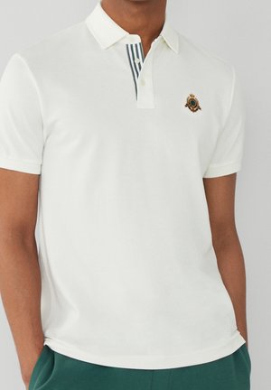 Man draagt een witte poloshirt met korte mouwen, met een gestreepte knoopsluiting en een geborduurd schildje op de linkerborst.