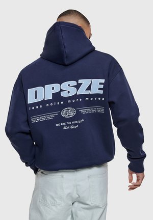 Mężczyzna w granatowej bluzie z kapturem z napisem i grafiką "DPSZE mniej hałasu więcej ruchu" na plecach, w połączeniu ze spodniami w jasnym kolorze.