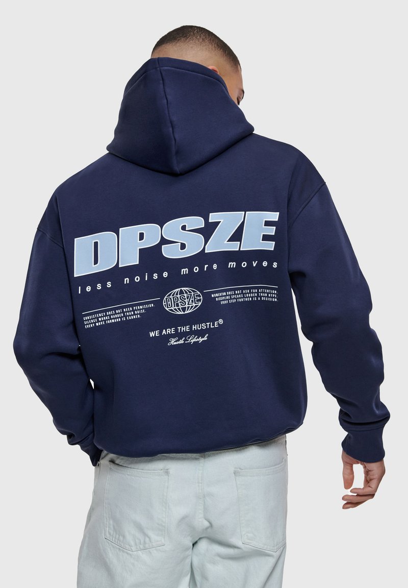 Homme portant un sweat à capuche bleu marine avec le texte et le graphisme « DPSZE less noise more moves » au dos, associé à un pantalon de couleur claire.