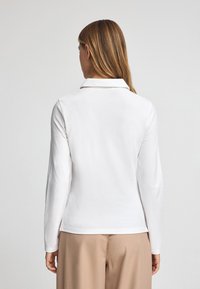 Långärmad, vit bomullspolo med krage. Har rak nederkant och avslappnad passform, bärs med beige byxor. Slät textur.