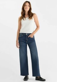 Donkerblauwe wijde jeans van denim, met een hoge taille, voorzakken en een nette afwerking. Gecombineerd met een witte tanktop en zwarte schoenen.