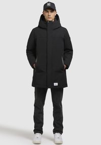 Sort polstret parka-jakke med hætte, lynlås foran, sidelommer og logomærke. Båret over mørke jeans og hvide sneakers.