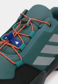 Sapato desportivo em azul-teal com atacadores elásticos laranja, fecho de atacadores azul e riscas cinzentas refletoras de lado.
