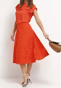 Röd silkblus med rynkad halsringning i kombination med en orange spetsmidikjol med blommiga utskärningar. Modellen håller en brun handväska.