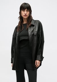 Camicia giacca in similpelle nera con vestibilità ampia, caratterizzata da tasche frontali e chiusura con bottoni, indossata sopra una maglia e pantaloni neri.