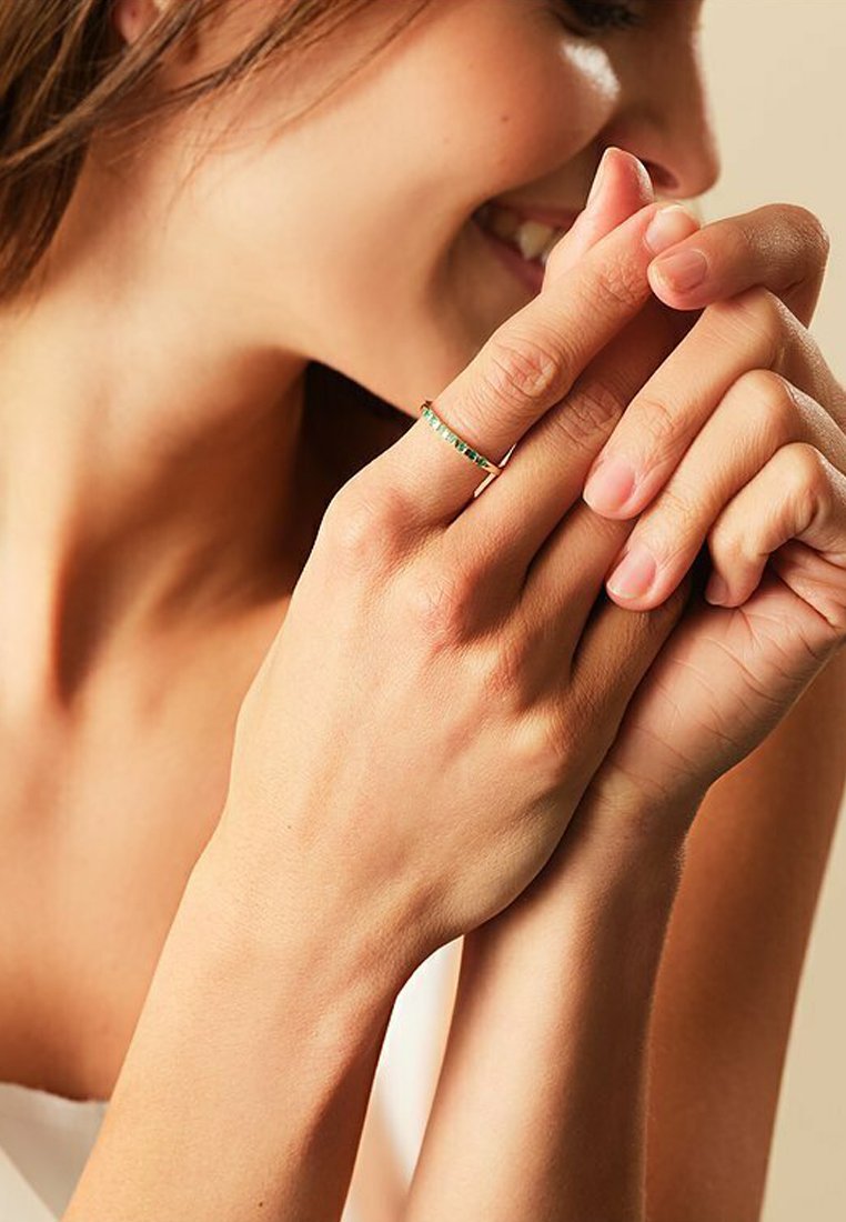 Goldring mit grünen Edelsteinen, der ein schlankes Design aufweist. Die Hand ist elegant positioniert und präsentiert den Ring auf der Haut.