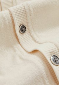 Tejido de color beige claro con una textura suave, que presenta detalles acanalados y un botón plateado. El material parece esponjoso y ligeramente texturizado.