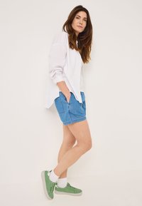 Camisa blanca de botones, pantalones cortos de mezclilla azul y zapatillas verdes con calcetines blancos. La camisa tiene las mangas arremangadas y un corte relajado.