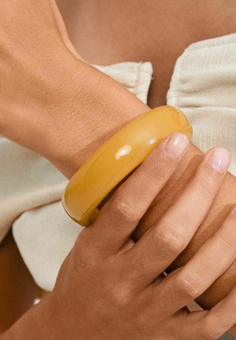Gul, glansig bangle-armband på ett handled. Banglen är slät och tjock, vilket står i kontrast till den texturerade huden och de naturliga naglarna som håller den.