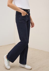 Jeans en denim bleu marine évasés avec une taille haute, cinq poches et des coutures contrastantes. Associés à des baskets blanches sur une surface texturée.