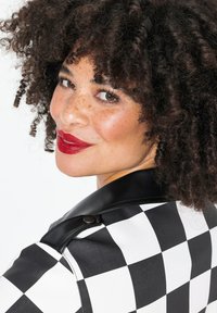 Veste à motif à carreaux noir et blanc avec un col en simili cuir, mettant en avant un accent en bouton. Le mannequin a les cheveux bouclés et les lèvres rouges.